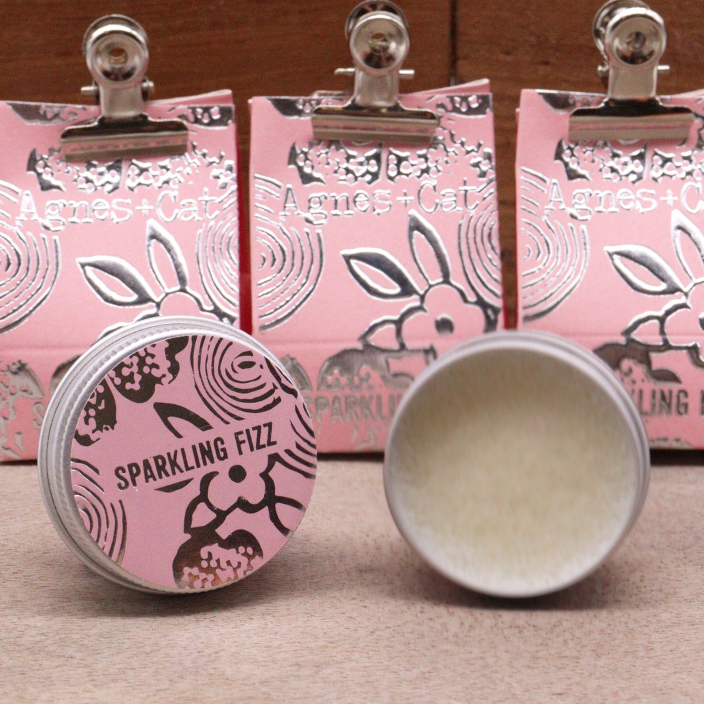 Cosmetic product with pink packaging and 'Sparkling Fizz' text, set against a wooden background.